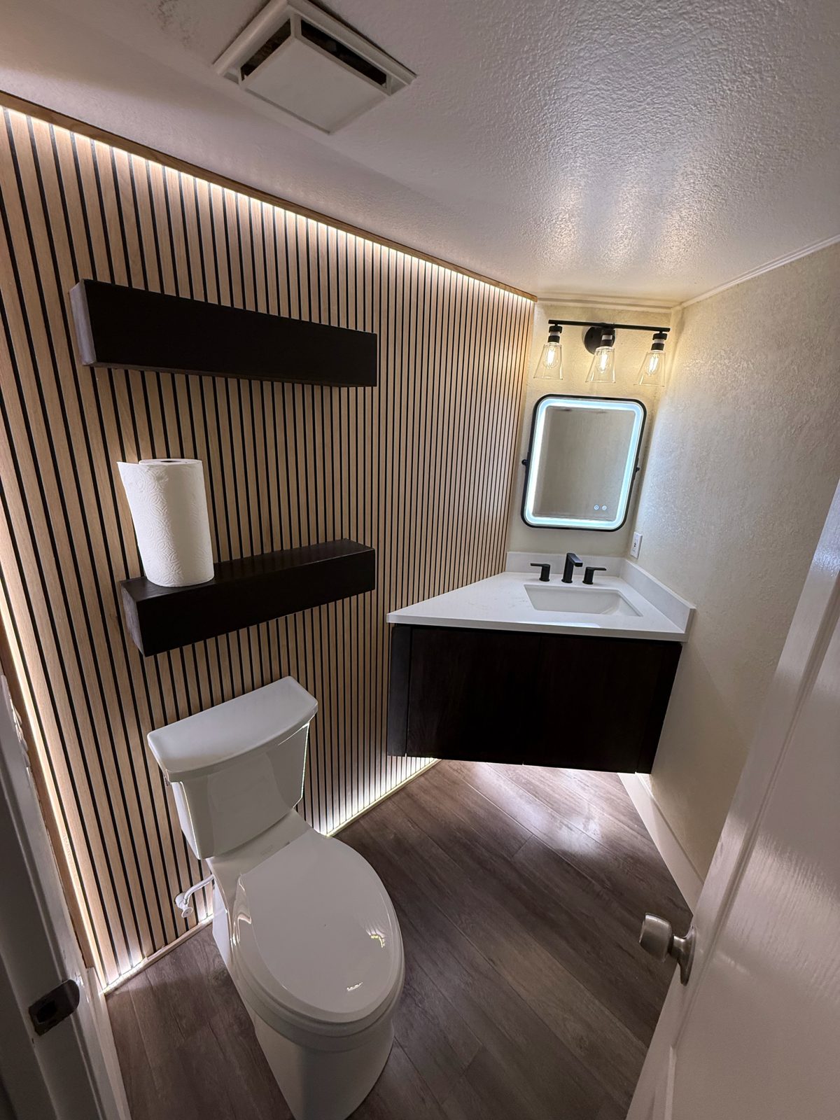 Powder room with a backlit wood slat wall, dark floating shelves and vanity, backlit LED mirror and black fixtures